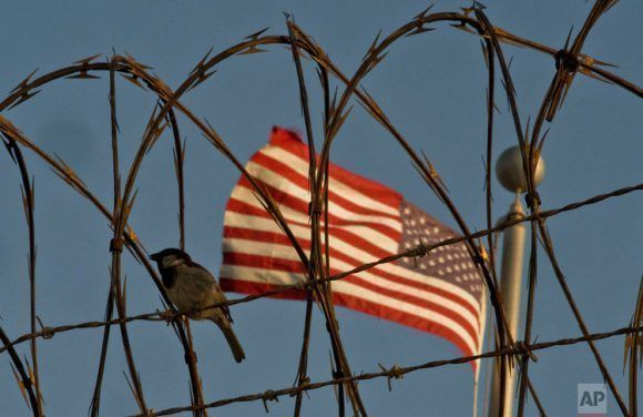 En esta foto del 5 de junio del 2018, revisada por oficiales del Ejército de Estados Unidos, un ave se posa sobre alambre espinado con la bandera estadounidense de fondo a las afueras de las instalaciones de detención del Campamento VI, en la Base Naval de Guantánamo. Foto: AP. (Tomada de Cubadebate)