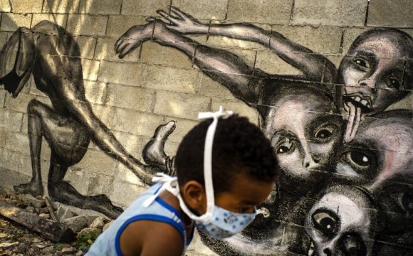 Un niño con una mascarilla para protegerse del coronavirus juega frente a un mural del artista Yuriel Peña titulado «Marcha hacia la oscuridad», en el patio de una casa en La Habana, Cuba, el viernes 10 de abril de 2020. Foto: AP /Ramón Espinosa