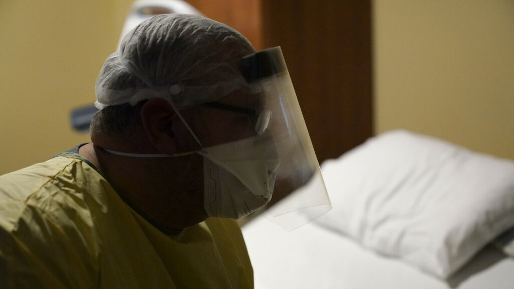 Un médico usa equipo de protección personal mientras se sienta en una cama para hablar con un paciente de COVID-19.Jeff Roberson / AP